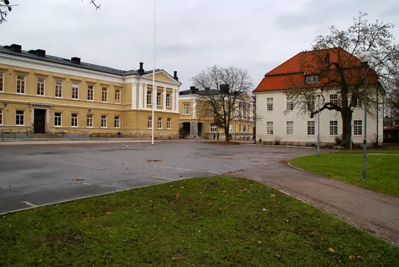 Höstbild som visar parkeringen vid Rudbeckianska gymnasiet. Här finns några kala träd, ett gult hus med svart tak och ett vitt hus med rött tak.
