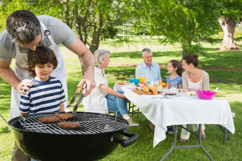 En man står och grillar med ett barn, i bakgrunden sitter en familj vid ett bord. 
