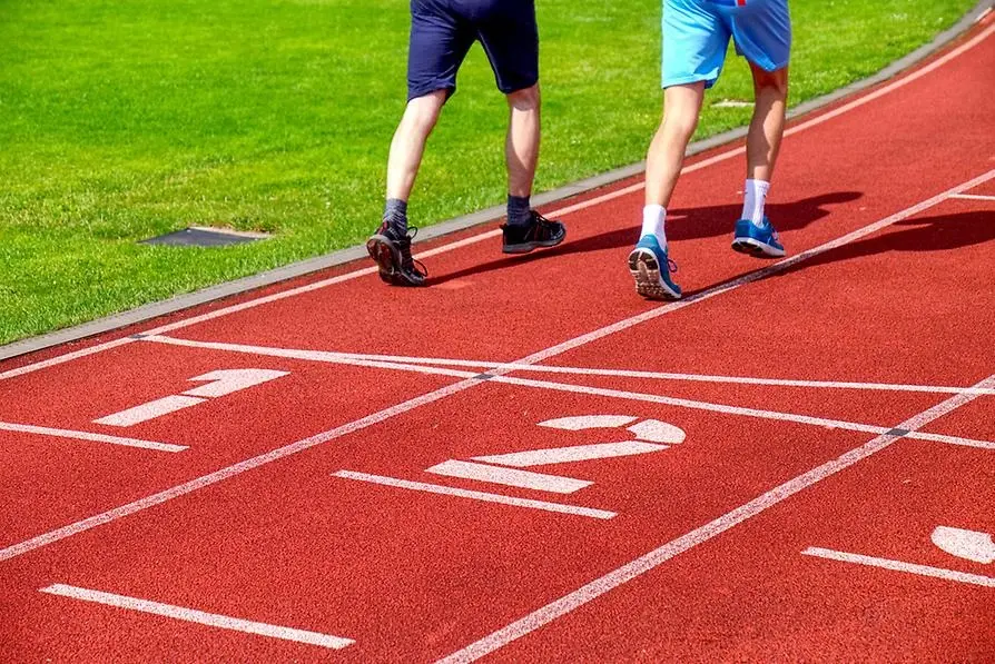 Benen på två personer som springer på en röd löparbana intill en gräsplan. Siffrorna 1 och 2 synd i nederkanten.